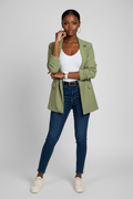 Woman wearing a green blazer, white top, blue jeans, and white sneakers on a gray background