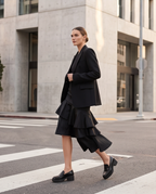 A woman walks across a city crosswalk wearing a black tailored blazer, a black tiered midi skirt, and black loafers, with a modern concrete building in the background.