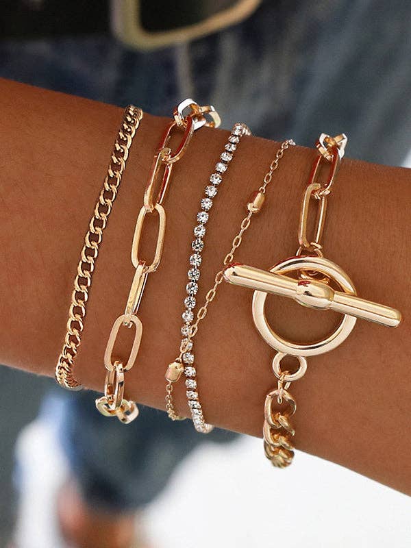 A close-up of a person's wrist wearing four layered gold-tone bracelets, including a curb chain, an elongated link chain, a rhinestone-studded chain, and a toggle ring bracelet.