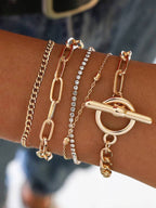 A close-up of a person's wrist wearing four layered gold-tone bracelets, including a curb chain, an elongated link chain, a rhinestone-studded chain, and a toggle ring bracelet.