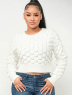 A woman wearing a white textured knit long-sleeve crop top sweater and blue jeans, standing against a plain white studio background.