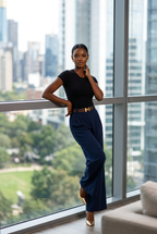 Woman in black top and blue pants standing by a large window with cityscape view